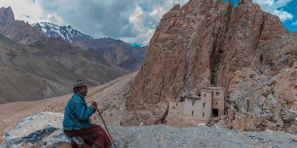 Man in traditional attire sitting on a rock in a mountainous landscape.