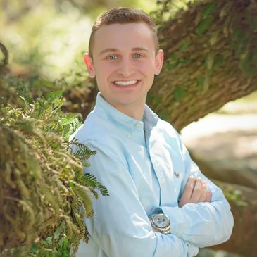 Young man smiling outdoors, leaning against a tree branch.