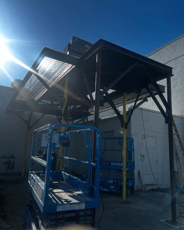 Blue scissor lifts under a metal platform with sun glare in an industrial area.