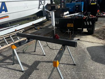 Metalworking setup with welded steel pieces on sawhorses outdoors near a boat trailer.