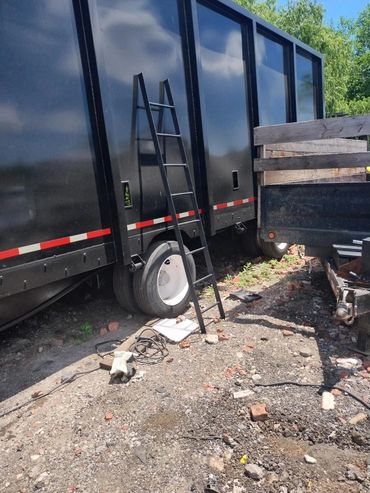 Black metal ladder leaning against a large black truck in a gravel area.
