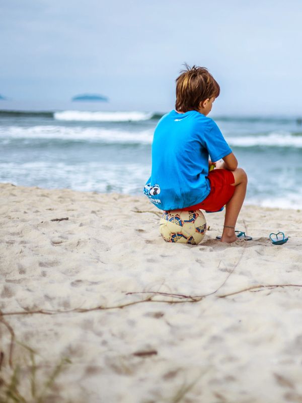 A boy in blue shirt and red shorts sits on a soccer ball at the beach.