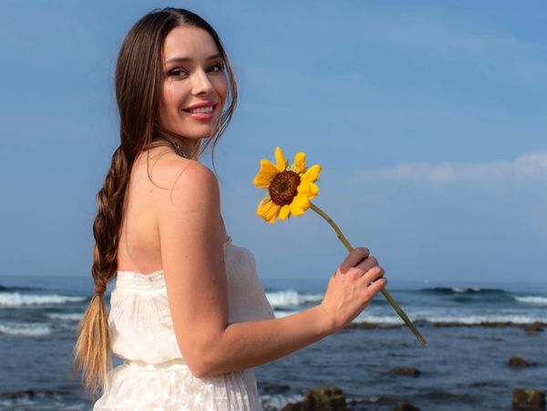 Woman in white dress holding a sunflower by the ocean, smiling.