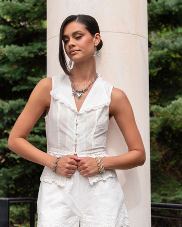 Woman in white sleeveless outfit wearing layered silver jewelry, standing by a column.