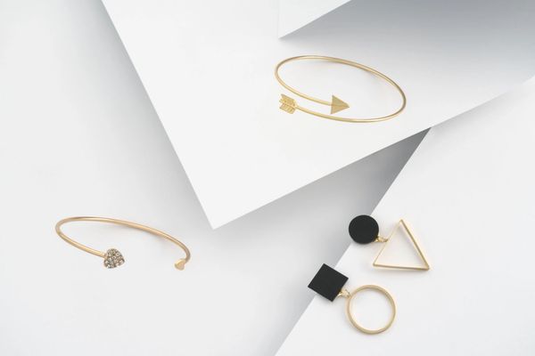 Gold jewelry on a geometric white background, featuring arrow and heart bangles, earrings.