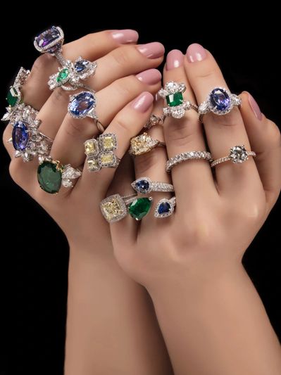 Hands adorned with multiple luxurious gemstone rings featuring emeralds, sapphires, and diamonds.
