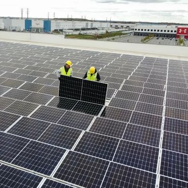 Workers installing solar panels on a large rooftop under cloudy skies.