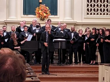 A portion of the group wearing black suits and gowns on stage while David addresses the audience