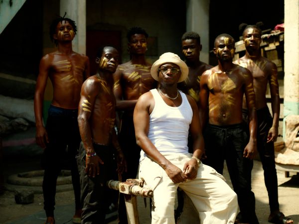 Group of men with yellow body paint, one seated wearing a hat and sunglasses.