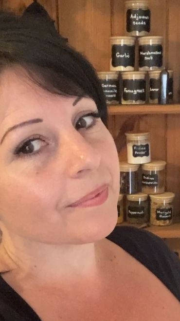 Woman smiling in a cozy kitchen with jars of spices and herbs behind her.