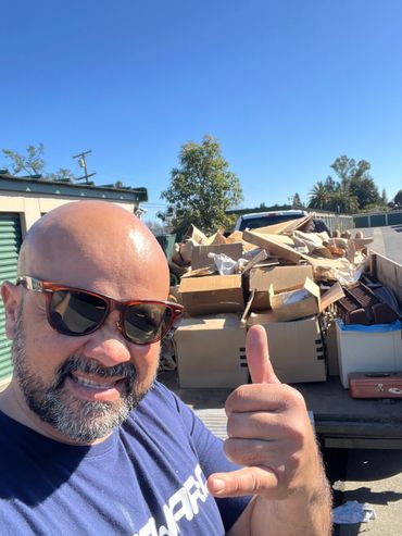 Man with sunglasses giving thumbs up in front of a truck loaded with cardboard boxes.