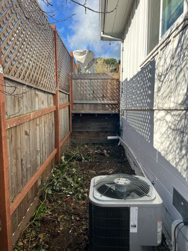 Narrow backyard space with wooden fence, air conditioning unit, and shadows from the lattice on the house wall.