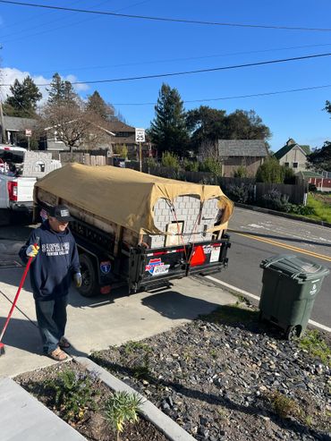 Man sweeping sidewalk next to a trailer covered with a tarp on a sunny day.