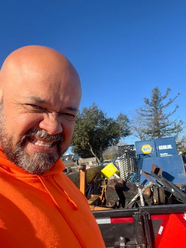 Man in orange hoodie smiles near a truck loaded with metal and tools under blue sky.