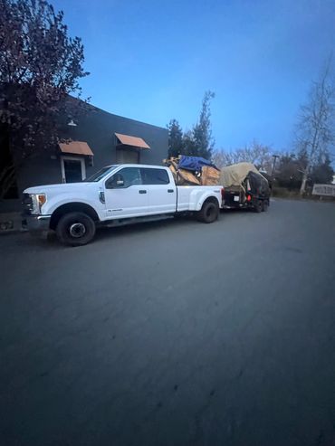 White pickup truck towing a trailer loaded with covered items on a street at dusk.