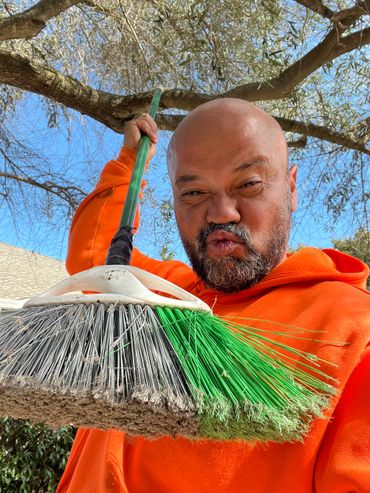Man in orange hoodie holding a broom with a playful facial expression outdoors.
