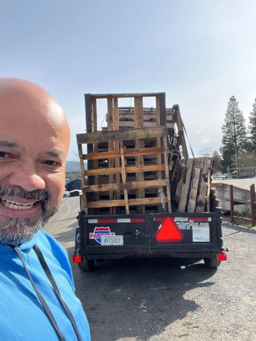 Man smiling beside a trailer loaded with wooden pallets on a sunny day.