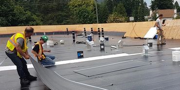 Workers installing roofing material on a flat roof under a cloudy sky.