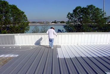 Person painting a metal roof white under clear blue sky.