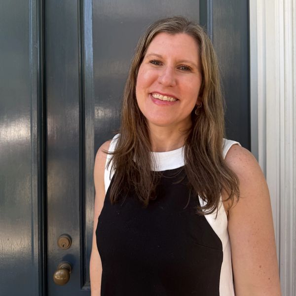 Smiling woman standing in front of a dark door wearing a black and white sleeveless top.