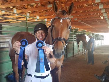 Smiling equestrian holding blue ribbons beside a horse in a stable.
