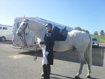 Smiling equestrian standing beside a white horse in a parking lot.