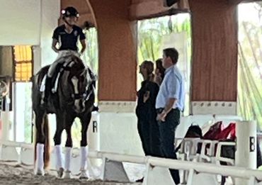 Rider on a horse receiving instructions from three observers indoors.