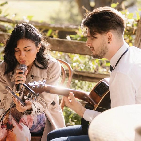 A woman sings into a microphone while a man plays guitar outdoors.