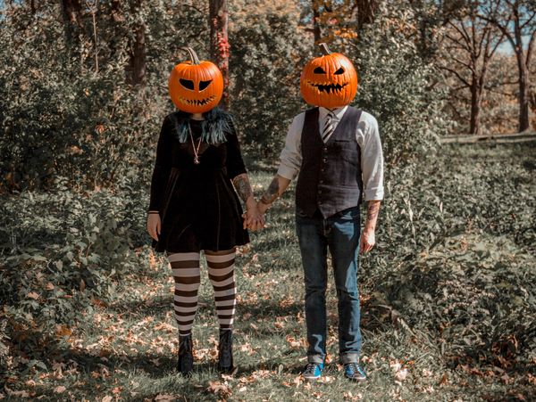 The Newlydeads wearing pumpkin heads standing in a forest holding hands. 