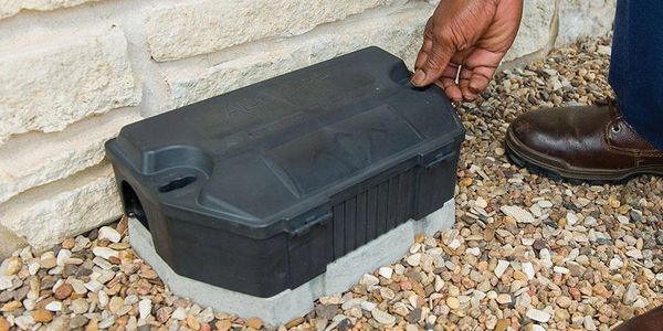Person opening a black plastic box on gravel beside a stone wall.