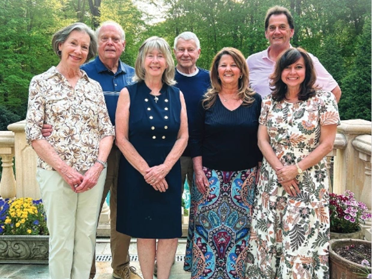 Seven adults smiling outdoors on a patio with floral decorations.