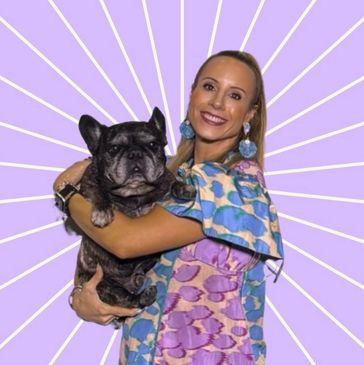 A woman in colorful dress holding a black French bulldog against a purple background.