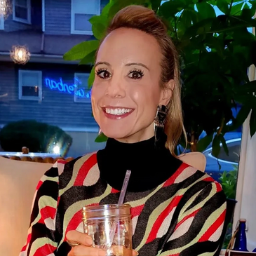 A smiling woman holding a drink in a colorful sweater.