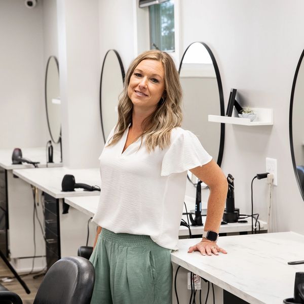 Smiling woman standing confidently in a modern hair salon with mirrors and chairs.