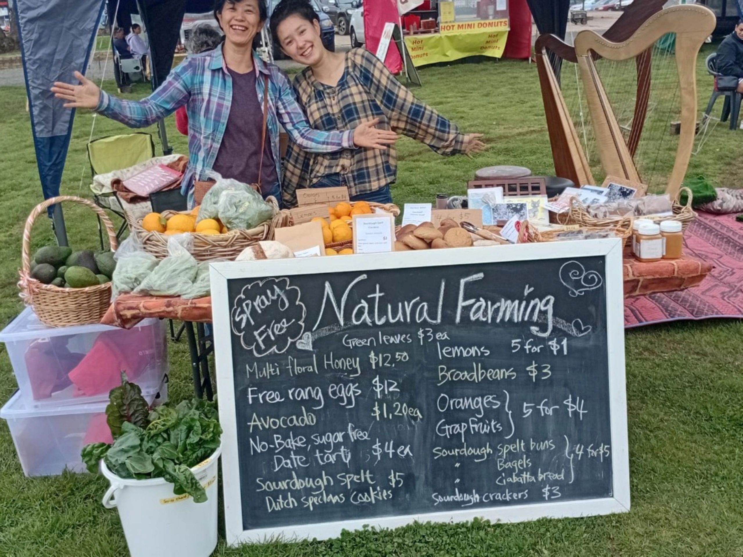 Two women at a natural farming market stall with fresh produce and baked goods.