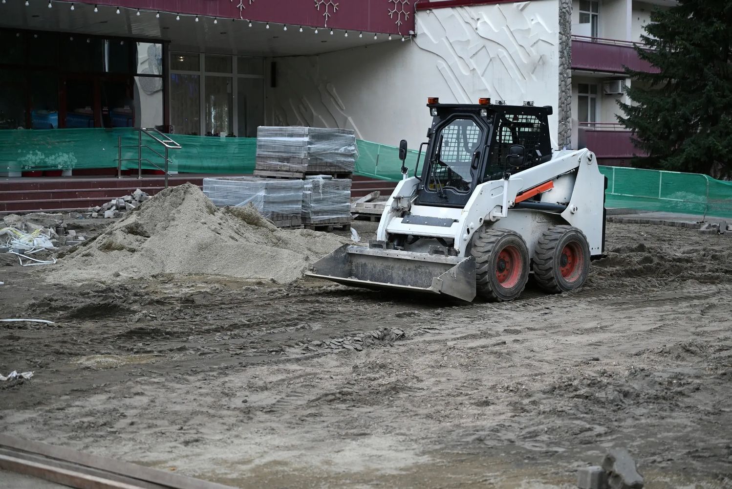 A skid-steer loader parked on a construction site with sand piles and stacked bricks.