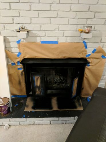 A fireplace being spray painted black with protective paper and blue painter's tape.