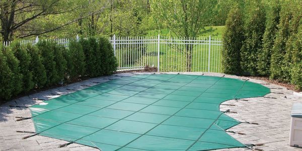 A backyard pool covered with a green safety cover, surrounded by trees and a white fence.
