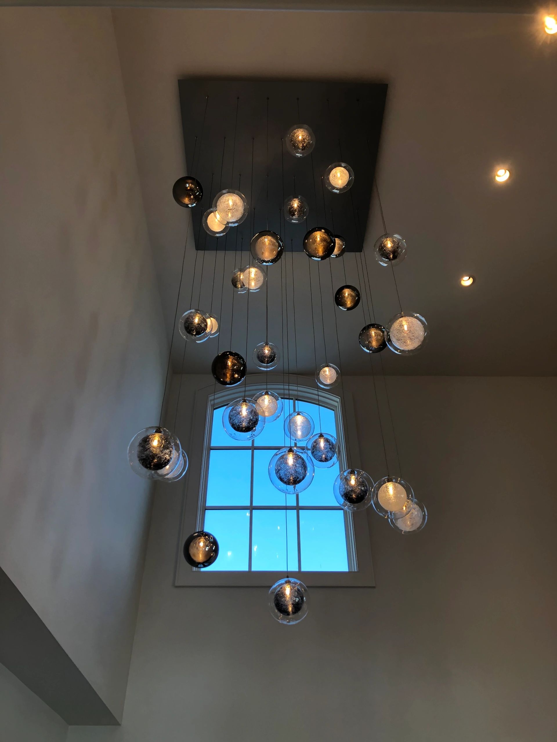 Modern hanging light fixture with multiple glass orbs illuminated near a large window.
