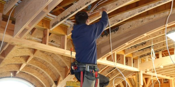Worker installing wiring on a wooden ceiling structure using a ladder.