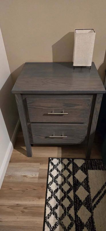 Gray wooden nightstand with a lamp on top and a patterned rug beside it.