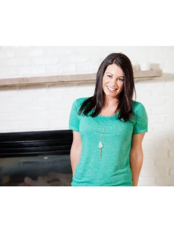 A smiling woman in a green shirt stands indoors in front of a white brick wall.