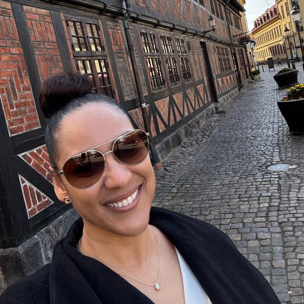Smiling woman in sunglasses takes a selfie on a charming cobblestone street with historic buildings.