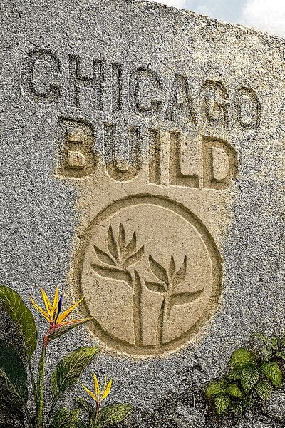 Engraved stone wall with 'CHICAGO BUILD' and plant logo, surrounded by tropical flowers.