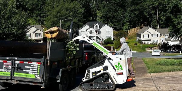 Worker loading a large log onto a truck using a mini loader in a suburban neighborhood.