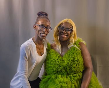 Two smiling women wearing glasses, one in a white cardigan and the other in a vibrant green ruffled dress.
