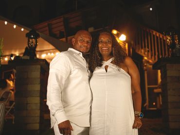 Couple dressed in white pose happily at a warmly lit nighttime event.