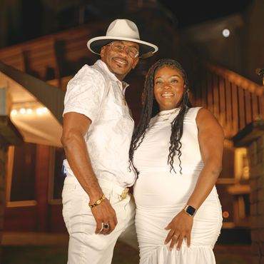 Couple dressed elegantly in white, smiling warmly under soft lighting.