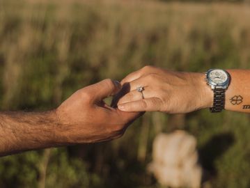 Two hands gently holding each other outdoors, one wearing a watch and ring.