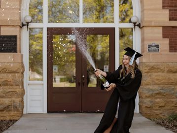 Graduate in cap and gown celebrating with champagne outside a building.